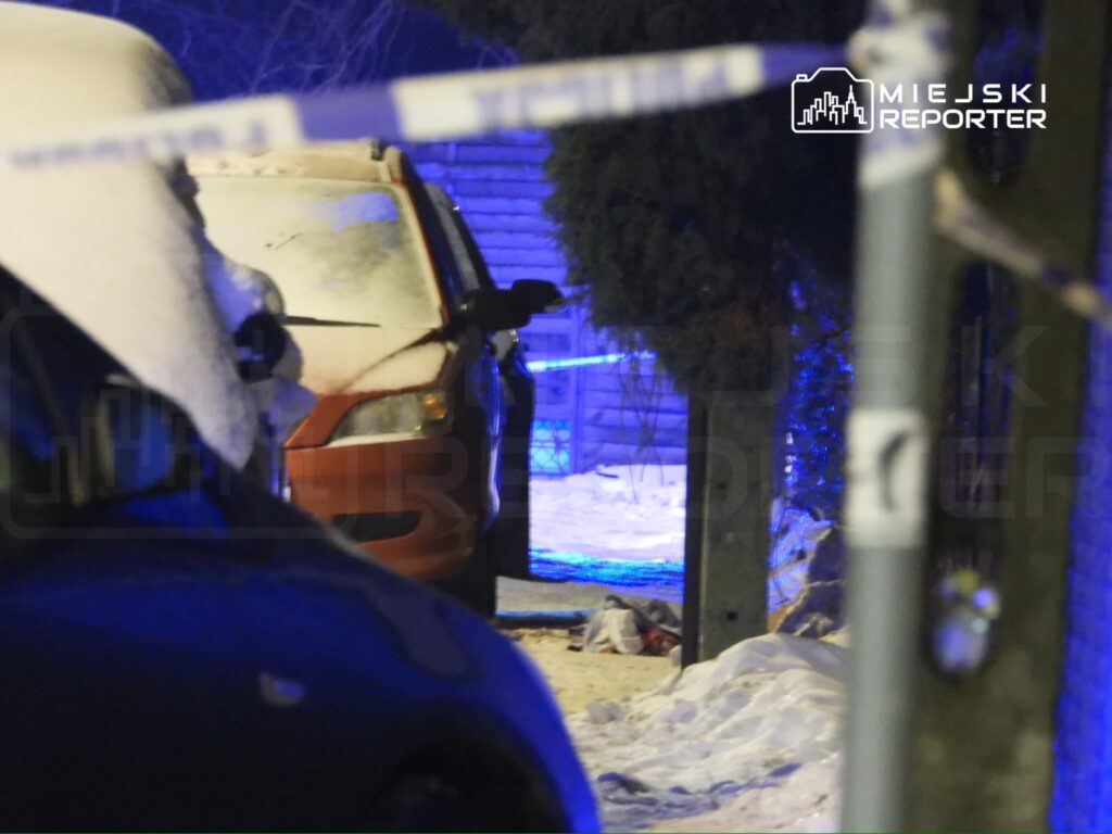 Czerwony samochód zaparkowany na śniegu, w tle niebieskie światła policyjne oraz taśma zabezpieczająca.