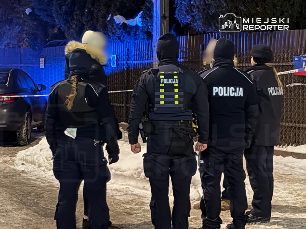 Grupa policjantów w odzieży ochronnej rozmawia w oświetlonym niebieskim świetle przy ogrodzeniu, w tle samochód na śniegu.