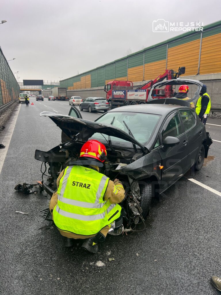 Strażak w odblaskowej kamizelce bada uszkodzenia samochodu osobowego na pasie drogi, obok ruchu tranzytowego.