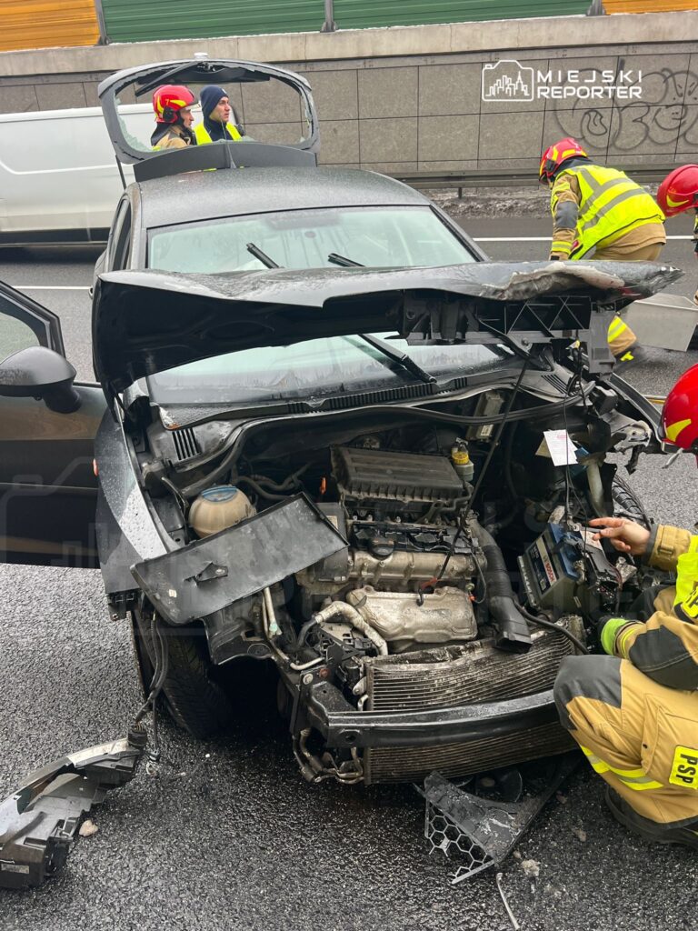 Na drodze stoi uszkodzony samochód osobowy, a strażacy w żółtych mundurach oceniają szkody.