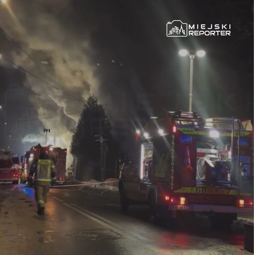 Strażak w mundurze zmierza w kierunku wozu strażackiego na oświetlonej ulicy, w tle widać dym unoszący się z pożaru.