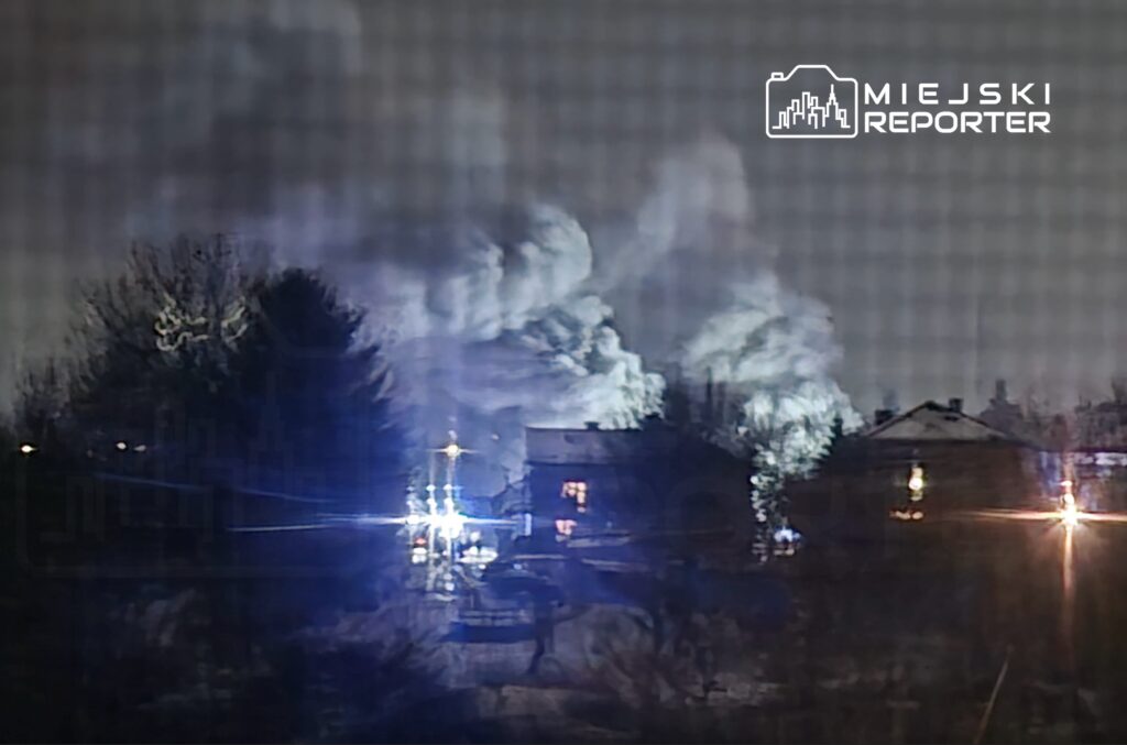 Zamglone niebo nocą z widocznymi smugami dymu oraz światłami pojazdów w tle.