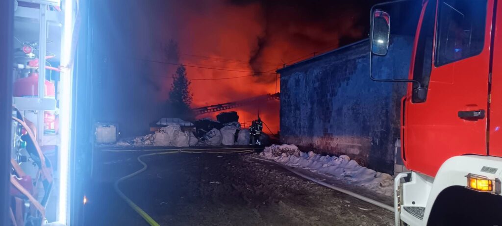 W tle płonący budynek, a z przodu strażak w pełnym umundurowaniu obok wozu strażackiego, w otoczeniu dymu i śniegu.