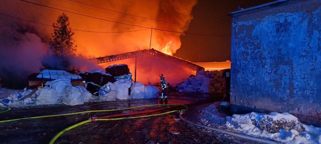 Strażak w specjalnym ubraniu gasi pożar budynku, który objęty jest dużymi płomieniami i dymem, w nocnej scenerii.