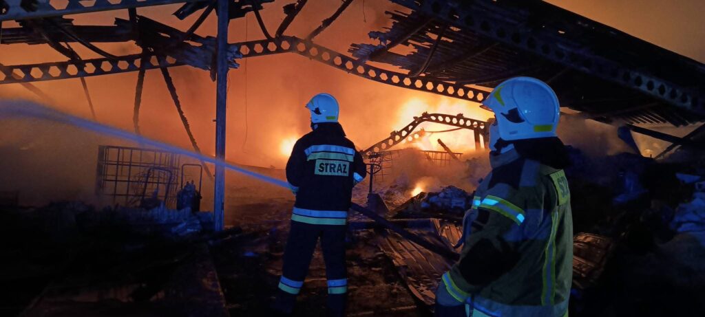 Strażacy w hełmach gaszą pożar zniszczonego budynku, w tle widoczne intensywne płomienie i dym.