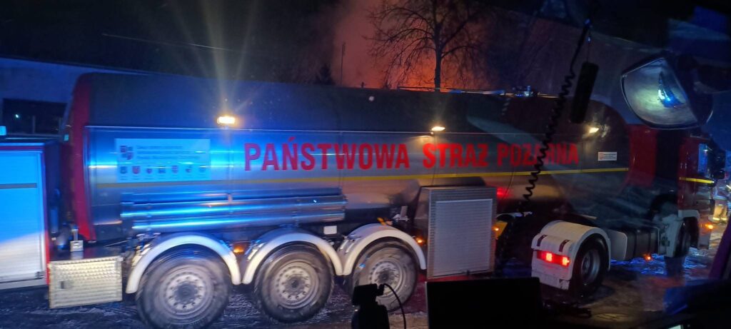 W nocy widoczny jest wóz strażacki z napisem "PAŃSTWOWA STRAŻ POŻARNA" w otoczeniu niebieskiego światła.