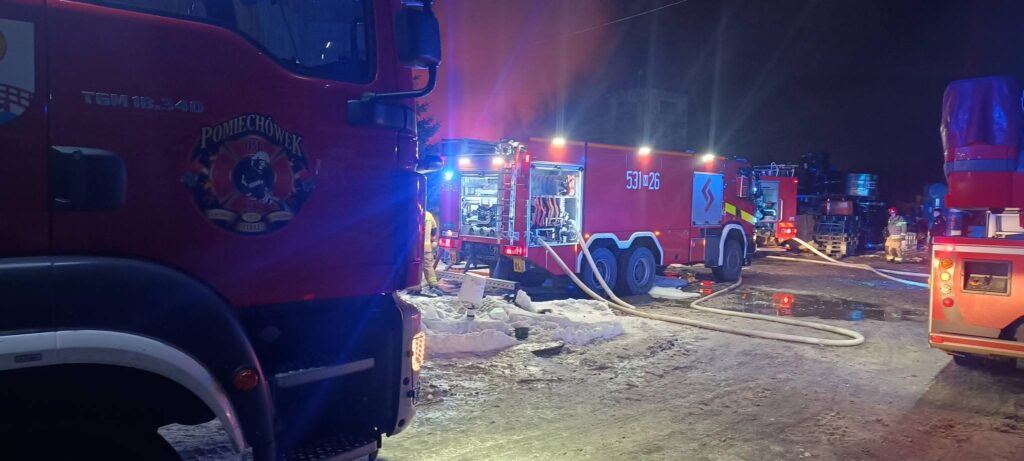 Czerwone wozy strażackie z włączonymi sygnałami świetlnymi, pracownicy w mundurach gaszą pożar w nocy.