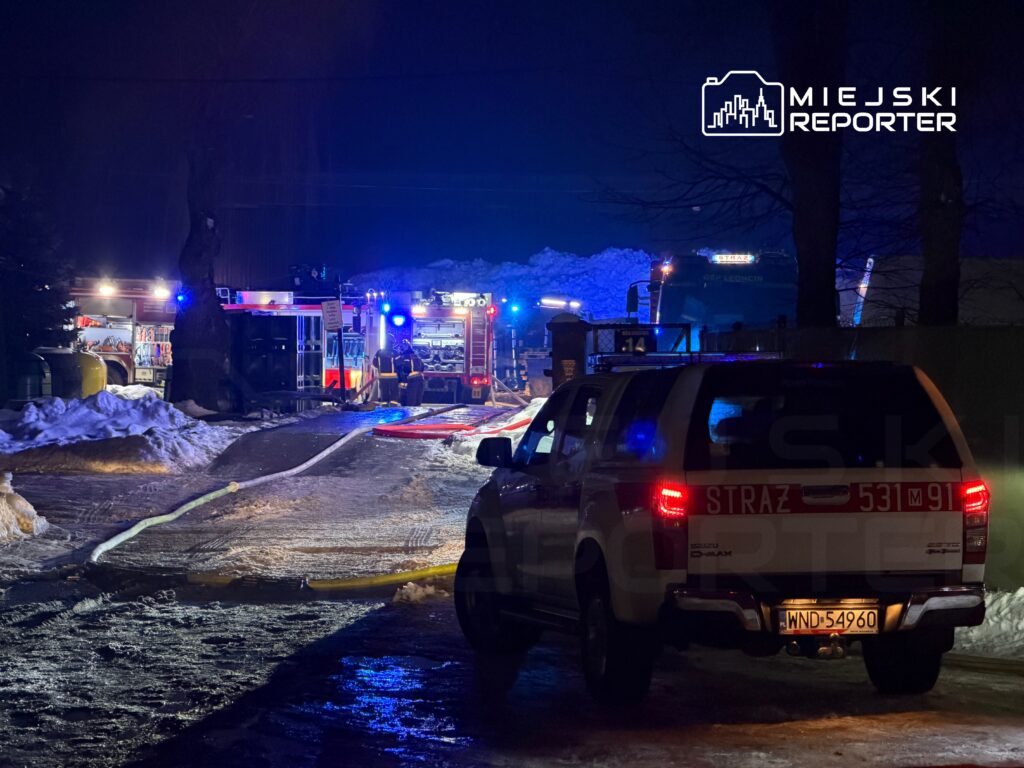 W nocy widać pojazdy straży pożarnej z włączonymi sygnalizacjami świetlnymi oraz węże strażackie na zaśnieżonej ulicy.