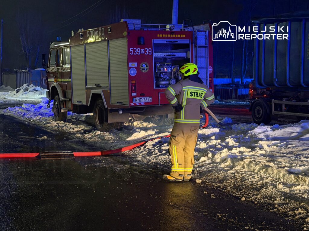 Strażak w żółtym hełmie podłącza węża gaśniczego do wozu strażackiego na ośnieżonej drodze nocą.