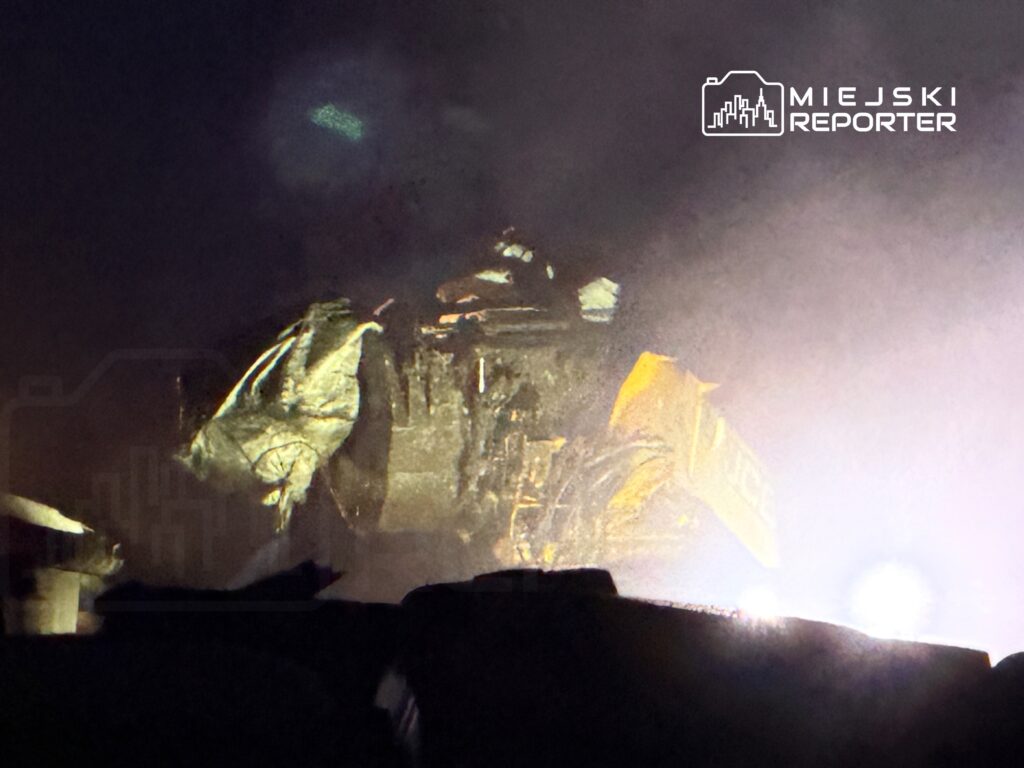 Maszyna budowlana pracująca w nocy, oświetlona przez reflektory, w tle widoczna mgła.