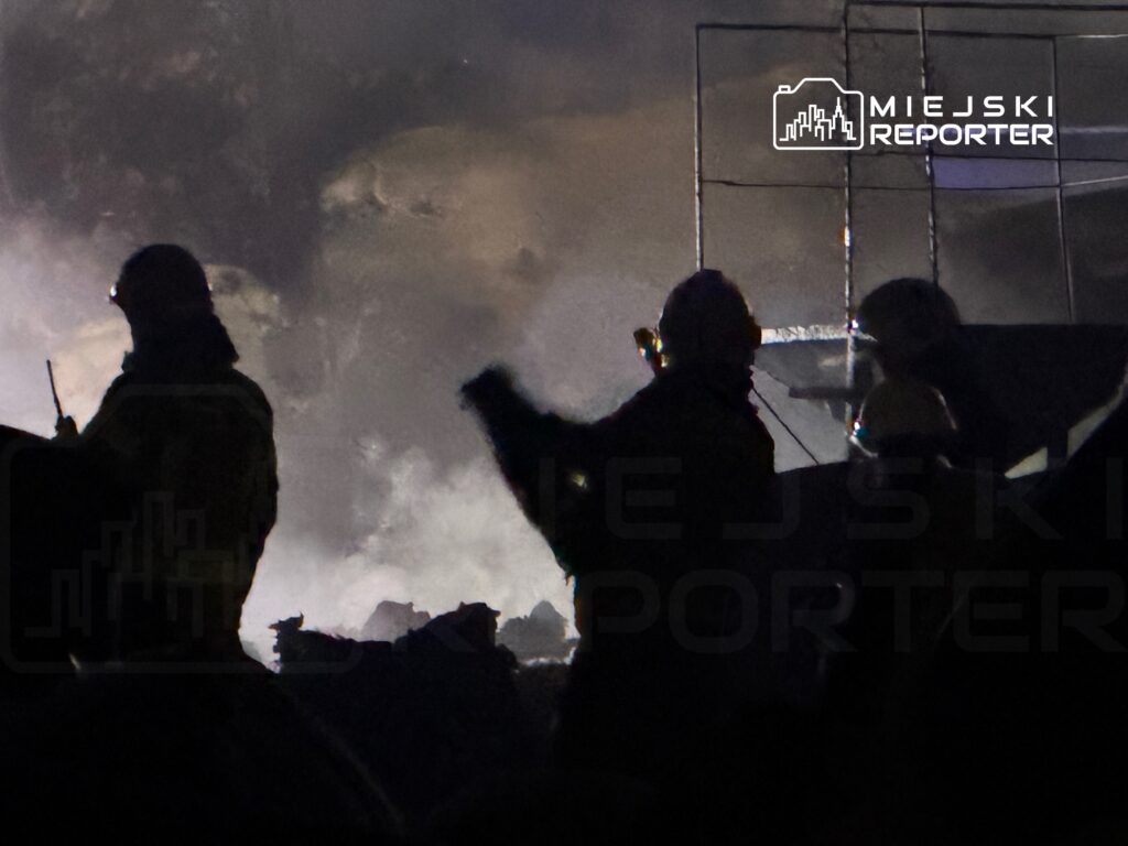 Grupa strażaków w hełmach i maskach, pracująca w zadymionym terenie podczas akcji gaśniczej.