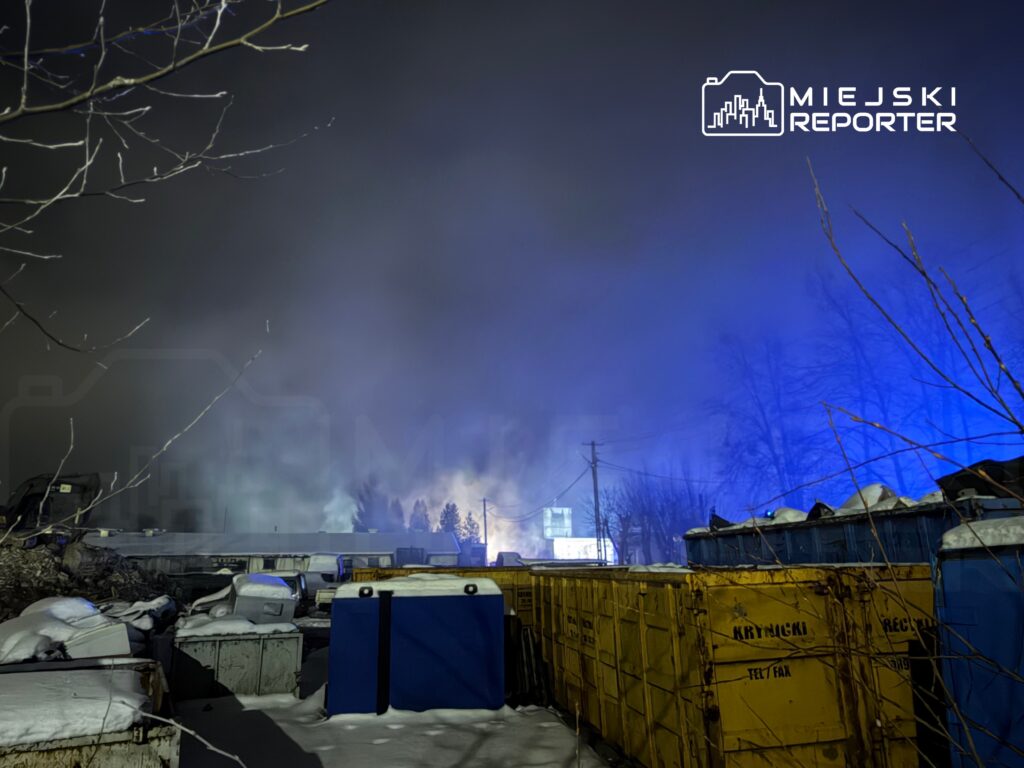 Na terenie przemysłowym w nocy widać dym i niebieskie światła, a w tle stoją kontenery i drzewa.
