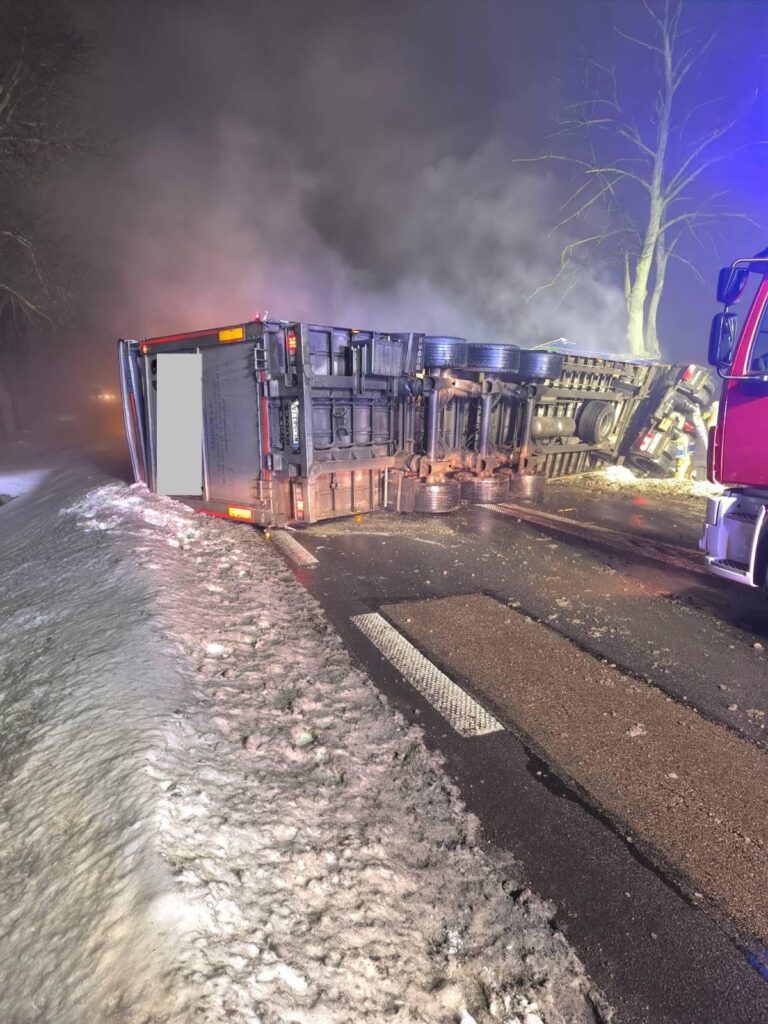 Cysterna przewrócona na drodze, otoczona dymem, obok widoczne ślady śniegu i interwencja straży pożarnej.