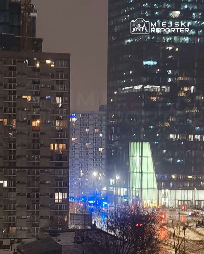 Nowoczesny biurowiec z przeszkloną fasadą w oświetlonej nocą miejskiej scenerii, w tle widoczne radiowozy.