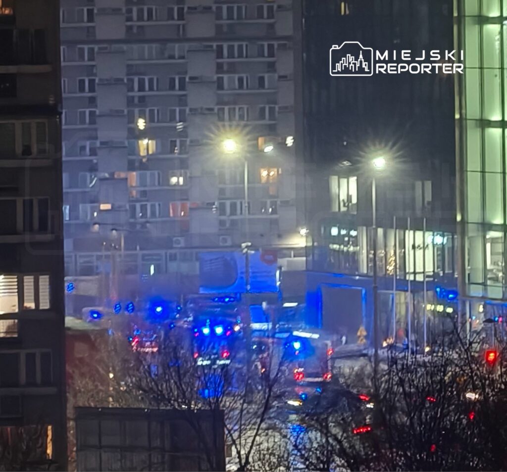 W nocy na ulicy widać liczne pojazdy służb ratunkowych z włączonymi sygnalizacjami świetlnymi oraz oświetlone budynki.