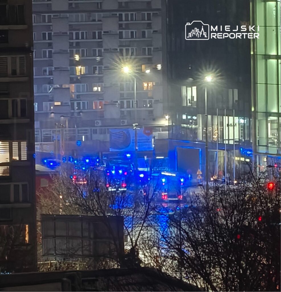 Niebieskie światła radiowozów oświetlają ulicę w nocy, w tle nowoczesny budynek z przeszkloną fasadą.