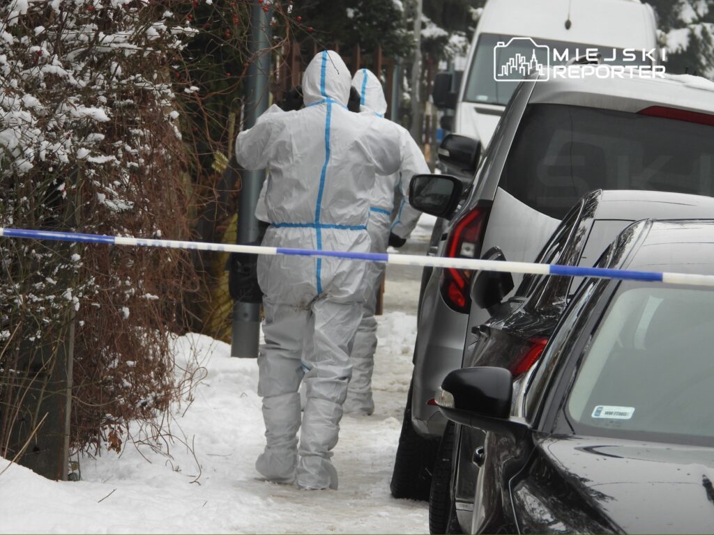 Dwaj mężczyźni w białych kombinezonach ochronnych prowadzą działania na zaśnieżonej ulicy, obok zaparkowanych samochodów.