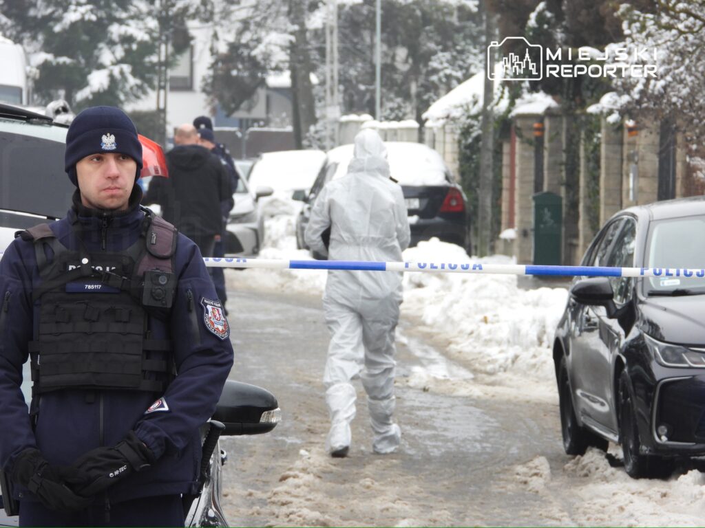 Policjant w mundurze stoi na zaśnieżonej ulicy, podczas gdy technik w białym kombinezonie bada teren za taśmą policyjną.