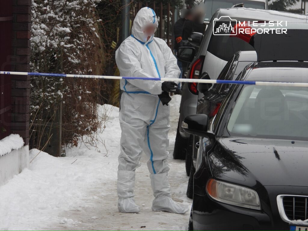 Funkcjonariusz w białym kombinezonie ochronnym zabezpiecza miejsce zdarzenia w zimowym otoczeniu, obok zaparkowanych samochodów.
