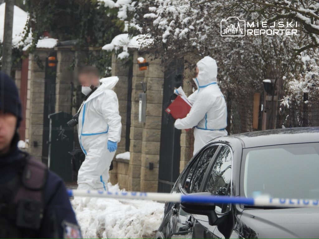 Grupa osób w białych kombinezonach ochronnych prowadzi działania w śnieżnym otoczeniu, obok zaparkowanego samochodu.