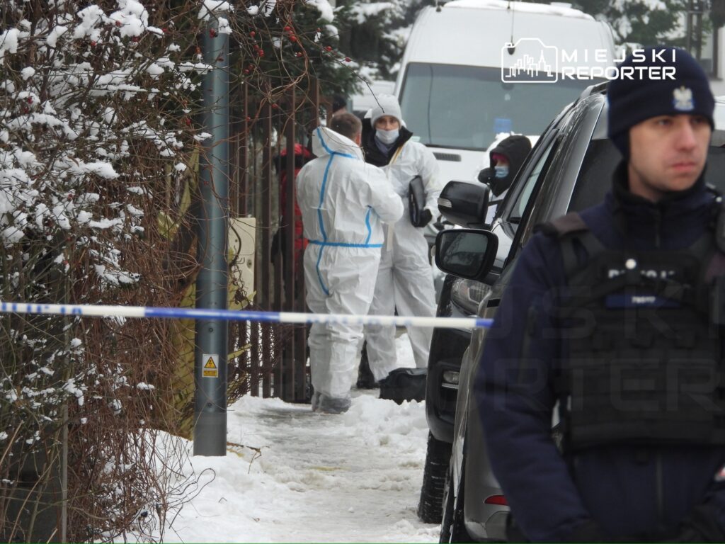 Grupa osób w białych kombinezonach bada miejsce zdarzenia w zaśnieżonym otoczeniu, a obok stoi policjant w kamizelce.