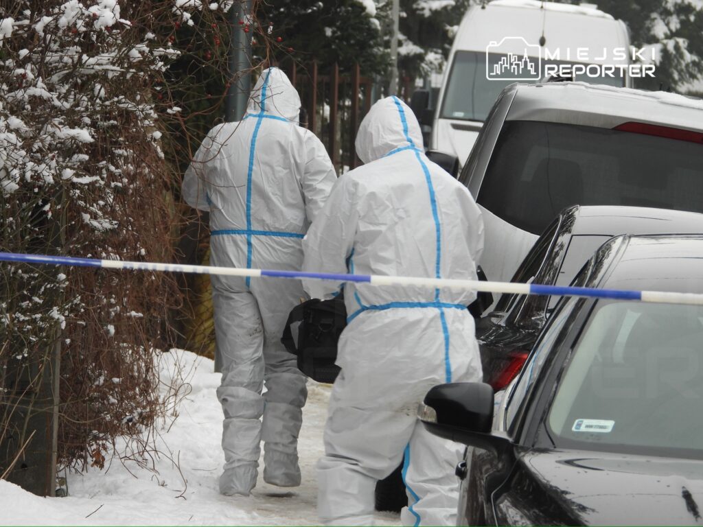 Dwaj mężczyźni w białych kombinezonach ochronnych przechodzą obok oznakowanego miejsca zdarzenia w zimowej scenerii.