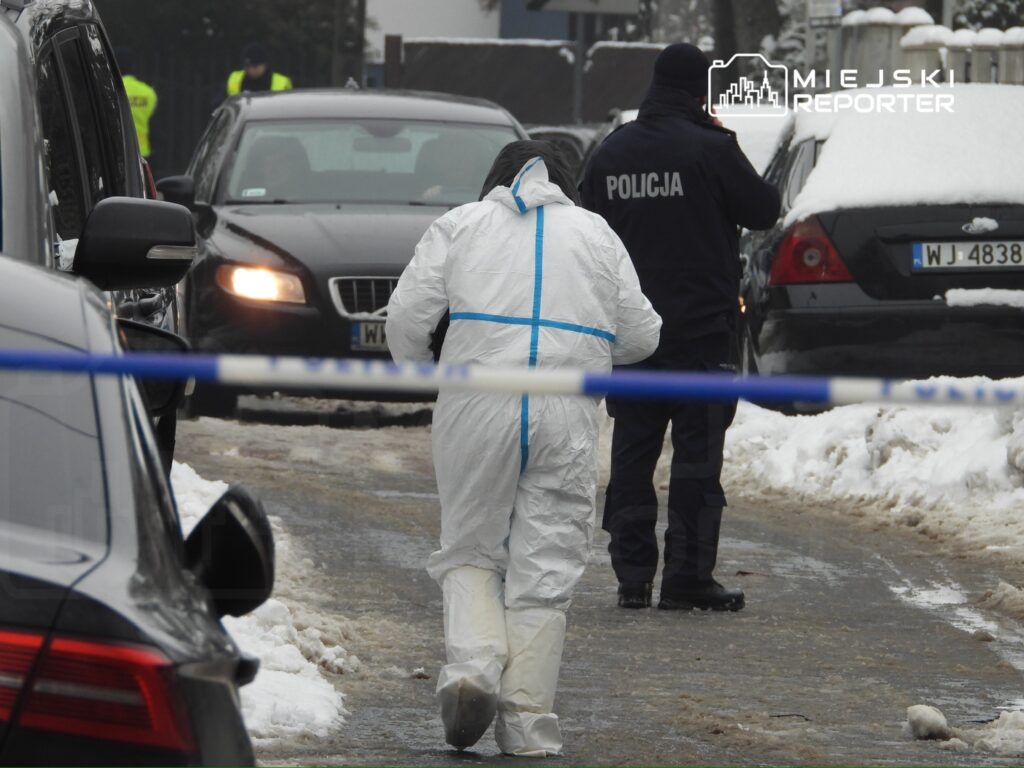 Osoba w białym kombinezonie ochronnym przemieszcza się wzdłuż miejsca zdarzenia, otoczonego policyjnym taśmą.