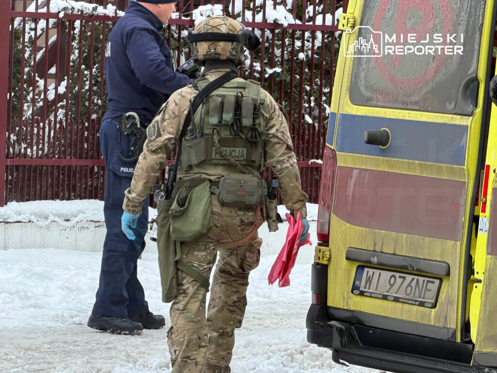 Funkcjonariusz policji rozmawia z żołnierzem w kamizelce taktycznej, obaj stoją obok karetki w zimowym otoczeniu.