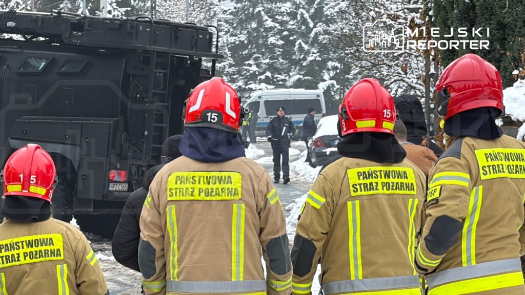 Grupa strażaków w hełmach obserwuje akcję ratunkową w zaśnieżonym terenie, obok stoi pojazd służbowy.