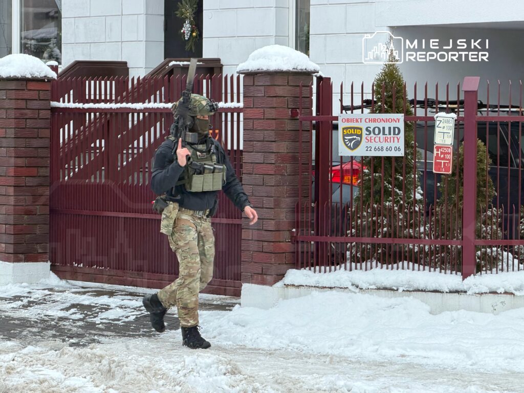 Mężczyzna w kamuflażu z bronią przechodzi obok ogrodzenia z tablicą "Obiekt chroniony Solid Security".