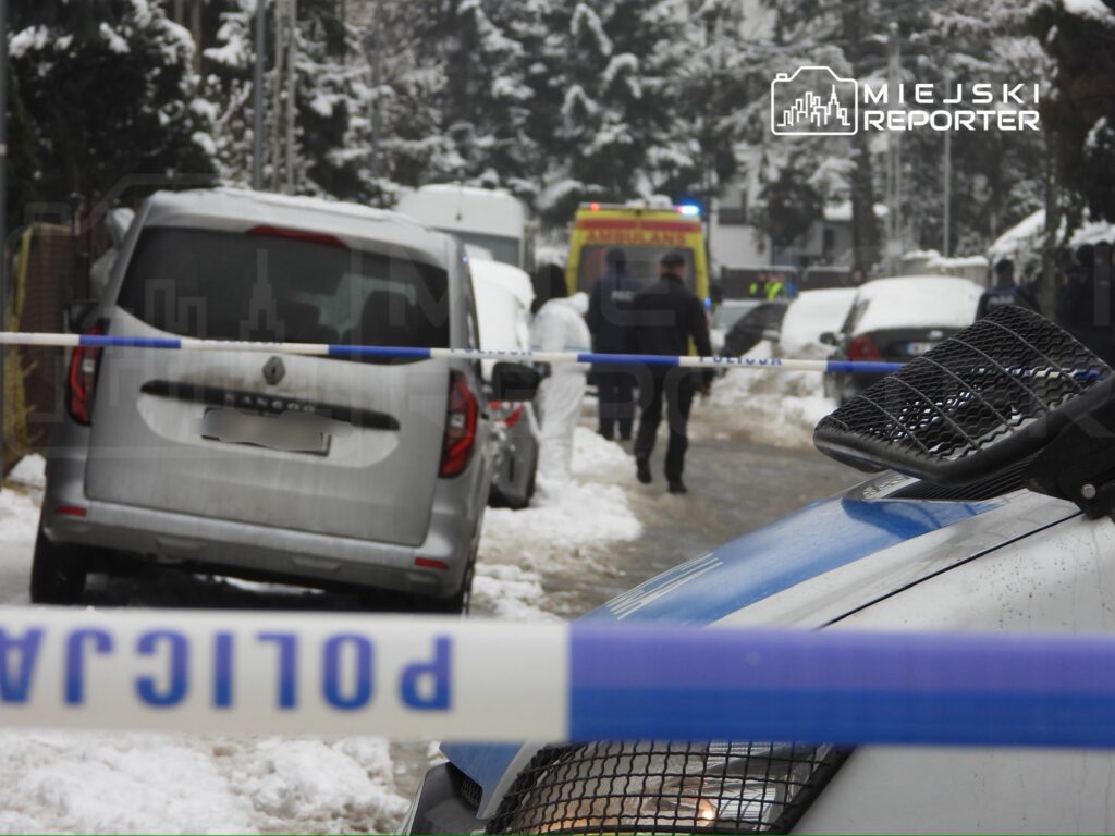 Na zaśnieżonej ulicy policyjny radiowóz stoi obok blokady, a funkcjonariusze prowadzą czynności dochodzeniowe.