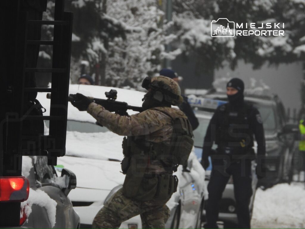Uzbrojony żołnierz w mundurze kamuflażowym celuje z bronią w stronę pojazdu, w otoczeniu zaśnieżonego krajobrazu.