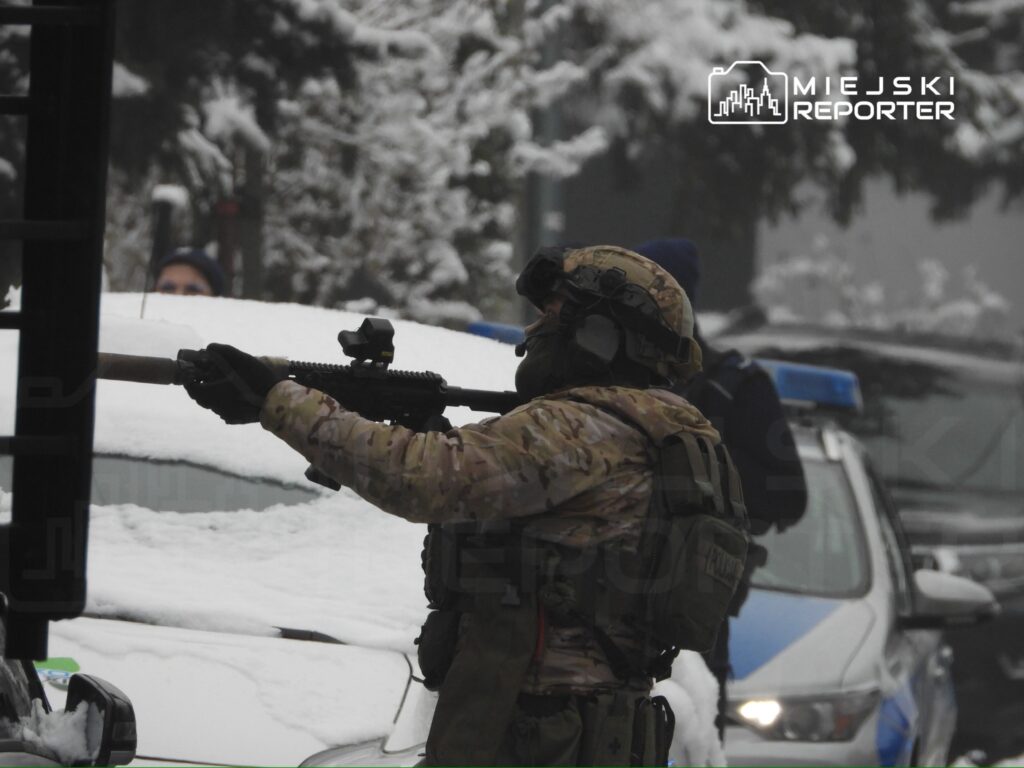 Żołnierz w kamuflażu trzyma karabin, obserwując okolicę w zaśnieżonym otoczeniu, obok radiowozów policyjnych.