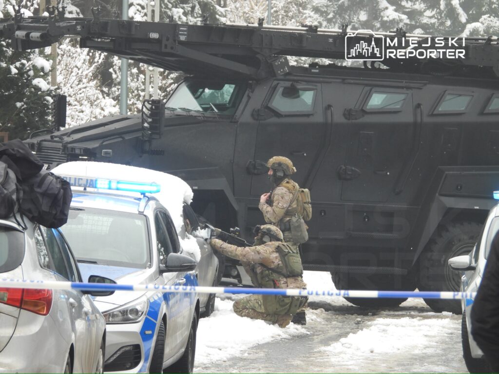 Żołnierz w kamizelce taktycznej przeszukuje samochód w pobliżu opancerzonego pojazdu, otoczonego przez policję.