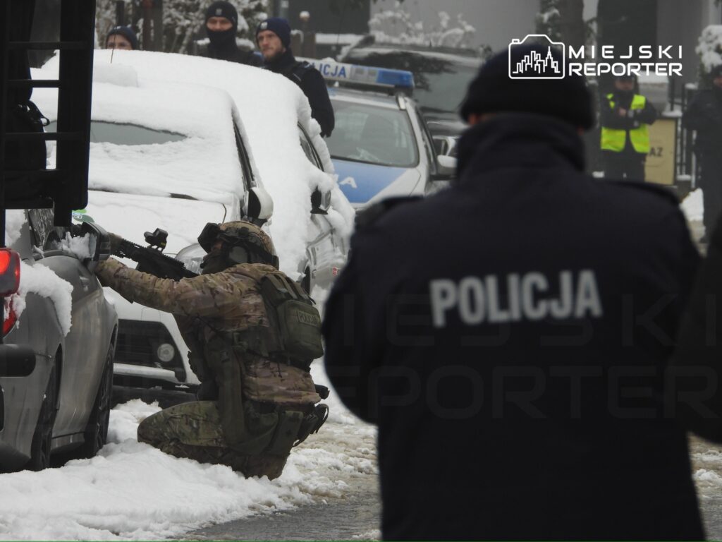 Żołnierz w mundurze z bronią klęczy obok samochodu przykrytego śniegiem, a w tle widać policjantów i pojazdy służbowe.