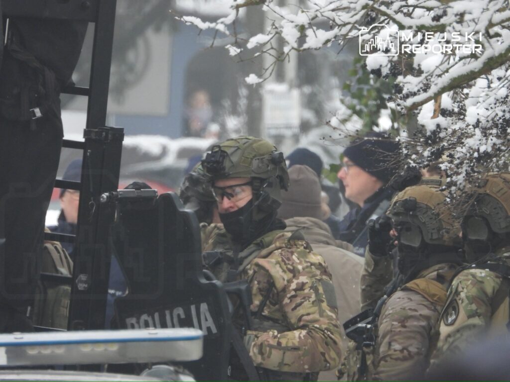 Grupa uzbrojonych policjantów w mundurach kamuflażowych przygotowuje się do akcji na zaśnieżonej ulicy.