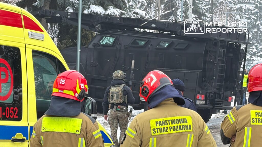Strażacy w hełmach obserwują działania policji przy opancerzonym pojeździe w zaśnieżonym otoczeniu.