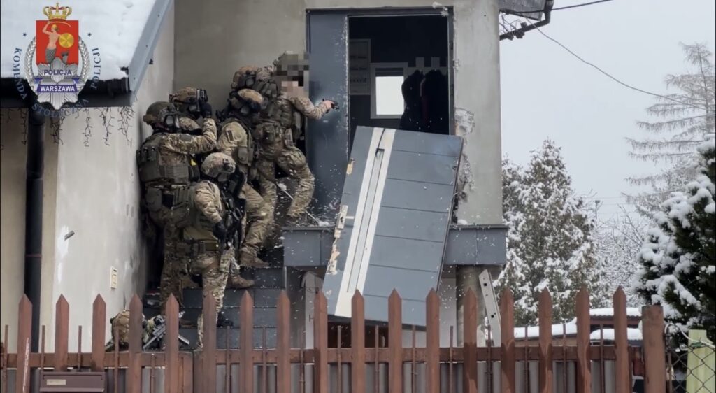 Grupa uzbrojonych funkcjonariuszy w kamuflażu wchodzi do budynku przez zburzone drzwi w zimowej scenerii.