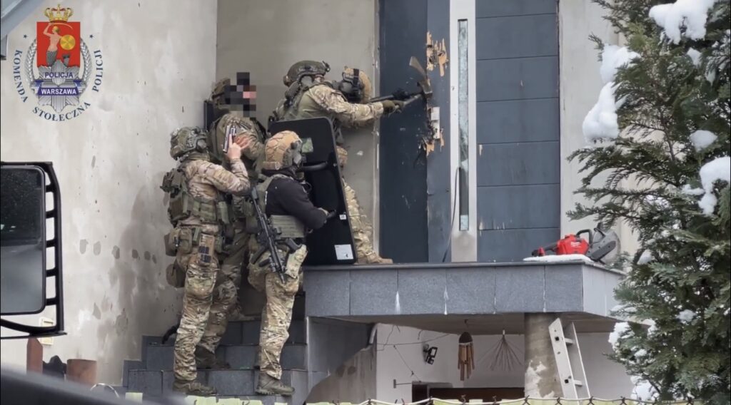 Grupa uzbrojonych funkcjonariuszy policji przygotowuje się do wejścia do budynku, używając narzędzi do wyważania drzwi.
