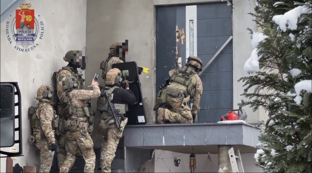 Grupa uzbrojonych funkcjonariuszy w kamizelkach taktycznych przygotowuje się do wejścia do budynku, przy drzwiach z widocznymi uszkodzeniami.