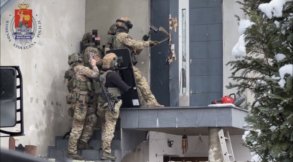 Czterech funkcjonariuszy w kamizelkach kuloodpornych używa młota do wyważania drzwi w zimowym otoczeniu.