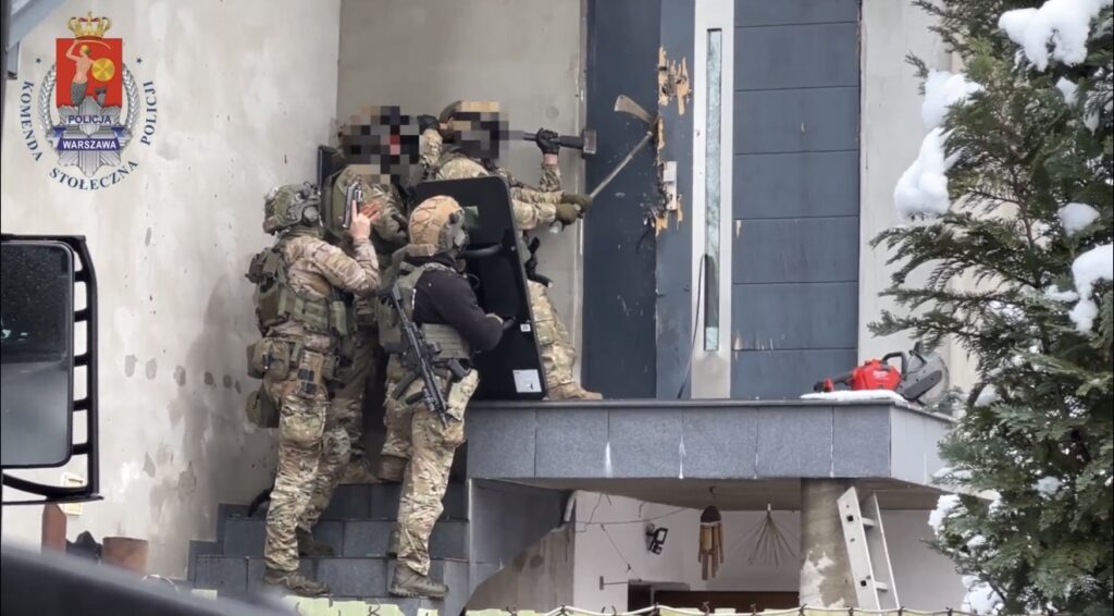 Grupa uzbrojonych policjantów w kamizelkach taktycznych atakuje drzwi wejściowe, używając siekiery.