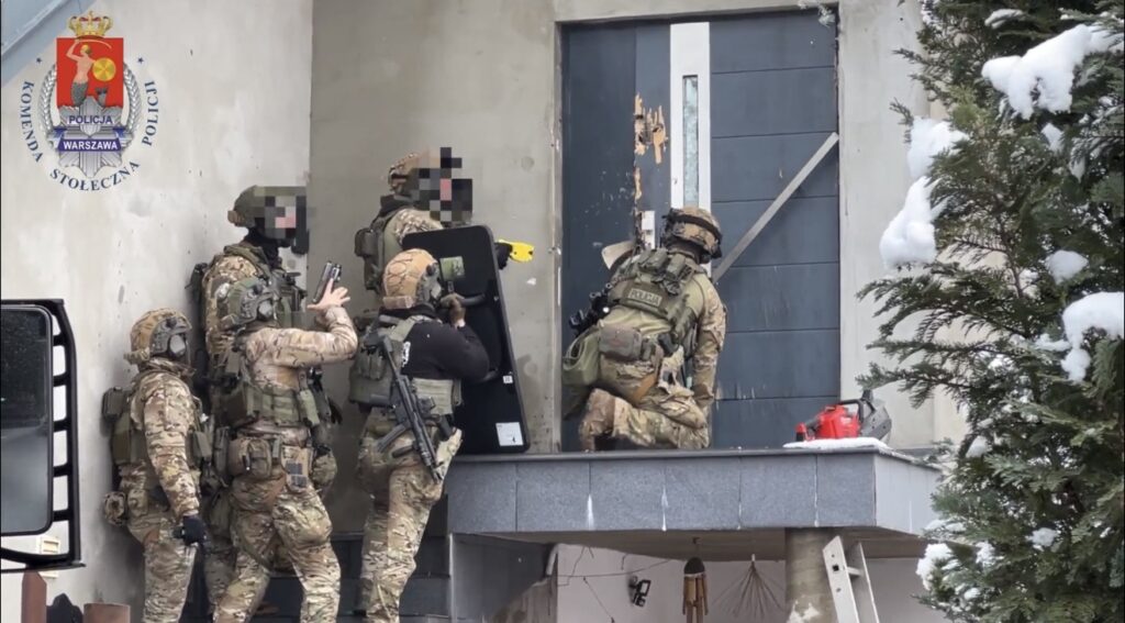 Grupa uzbrojonych policjantów w kamizelkach kuloodpornych przygotowuje się do wejścia do budynku, korzystając z tarczy ochronnej.