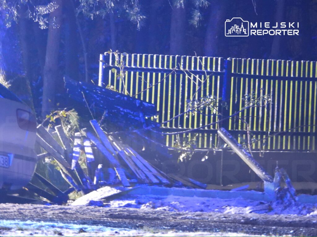 Złamany płot leży na ziemi obok białego samochodu, w tle widać drzewa oświetlone niebieskim światłem.