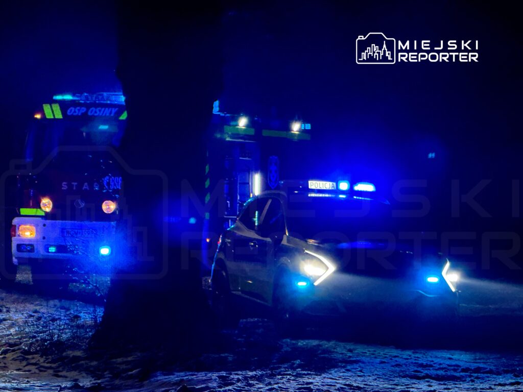 W nocy pojazdy straży pożarnej i policji z włączonymi sygnałami świetlnymi stoją na zaśnieżonej drodze.