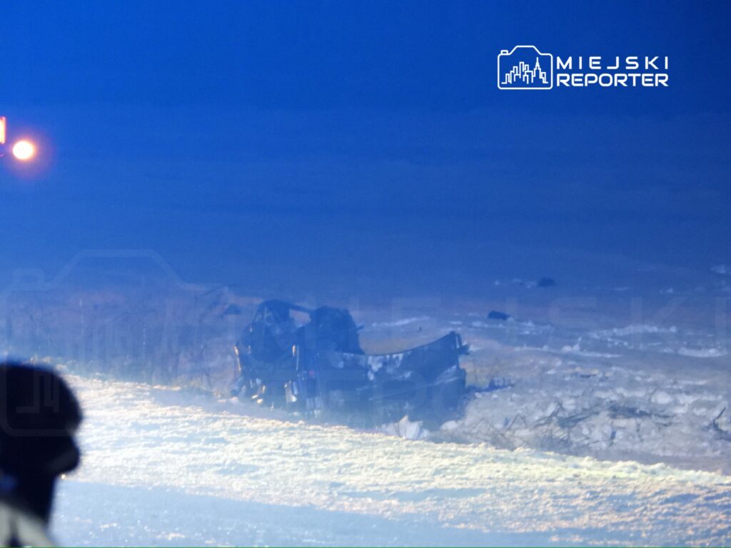 Zniszczony pojazd leży na poboczu drogi, otoczony śniegiem i mrokiem nocnego krajobrazu.