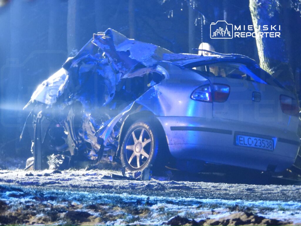 Zniszczony samochód osobowy leży na poboczu drogi w lesie, oświetlony niebieskimi światłami służb ratunkowych.
