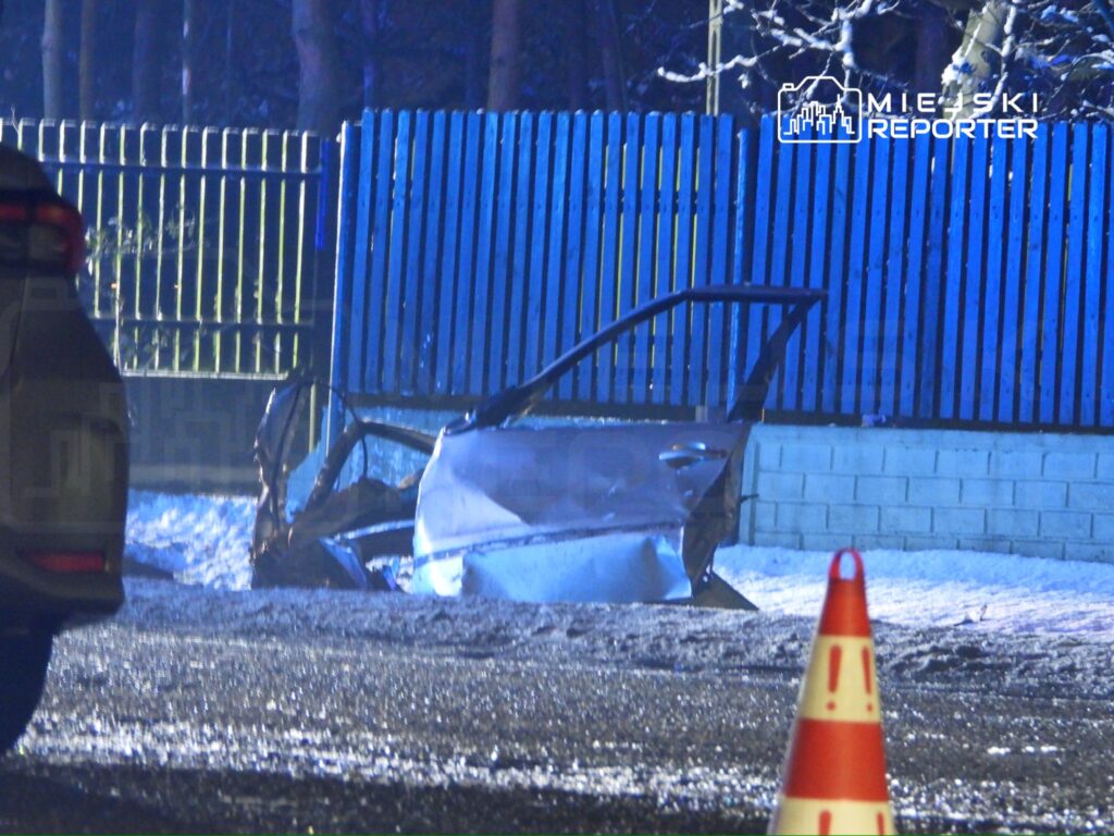 Zniszczona karoseria samochodu leży na poboczu drogi, obok oznaczenia ostrzegawczego w nocy.