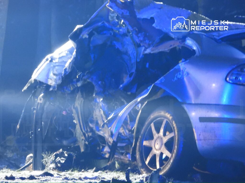 Zniszczony samochód osobowy leży na poboczu drogi, oświetlony niebieskimi światłami policyjnymi.