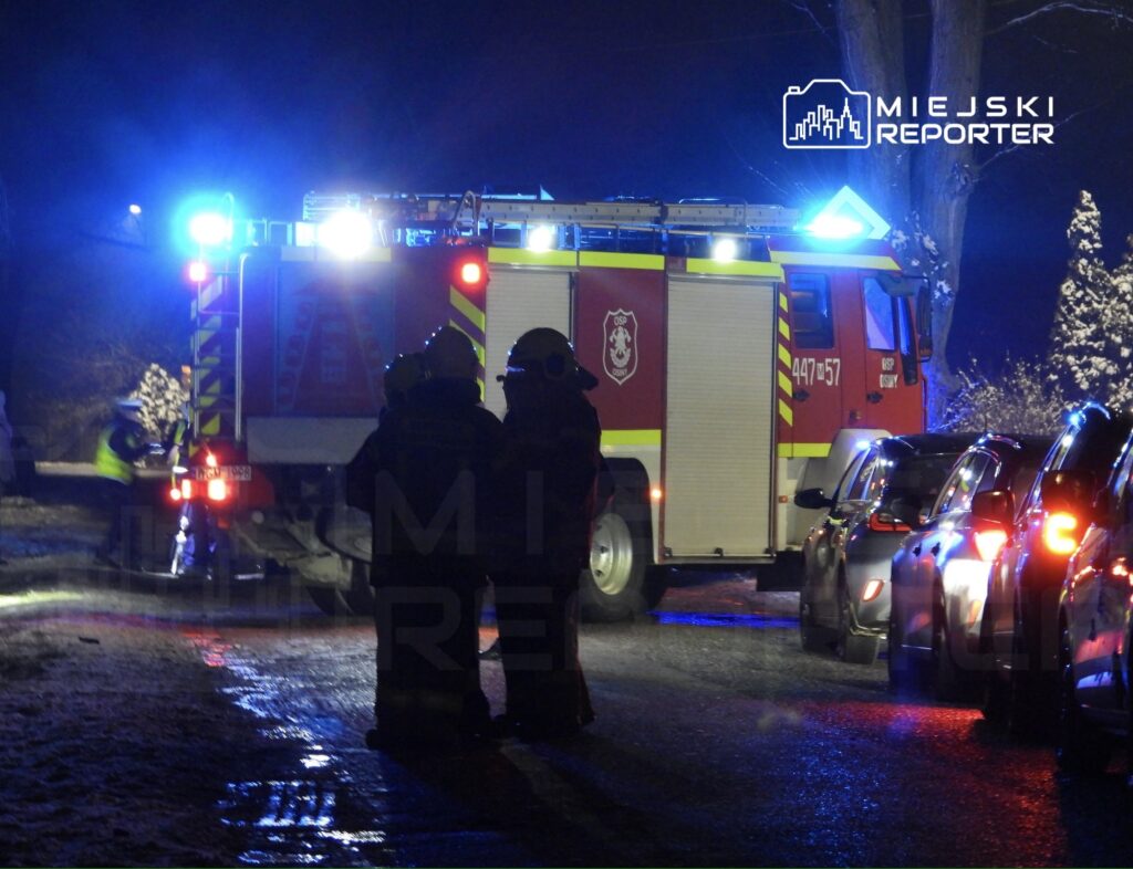 Strażacy w hełmach obserwują akcję ratunkową przy wozie strażackim z włączonymi sygnałami świetlnymi.